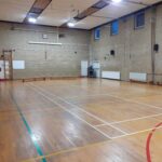 Empty indoor gym with brick walls, wooden floor, and climbing frame along the left wall; various court lines visible on the floor.