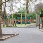 Children's playground with wooden climbing frames, tires and a slide, surrounded by a fence and trees.