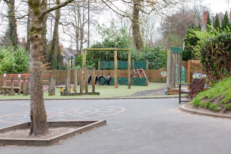 Children's playground with wooden climbing frames, tires and a slide, surrounded by a fence and trees.