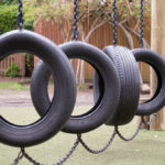 Tire swings suspended by chains in a playground with a wooden post and green turf beneath.