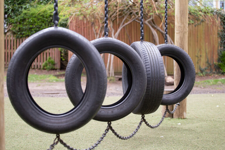 Tire swings suspended by chains in a playground with a wooden post and green turf beneath.