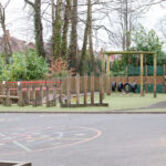 Children's outdoor playground with climbing wall, tires, and wooden balance structures, enclosed by a fence and surrounded by trees.