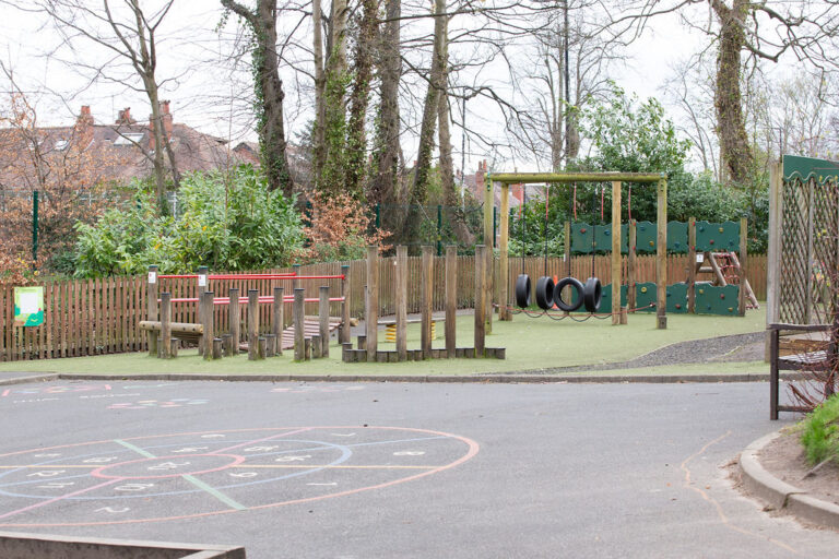 Children's outdoor playground with climbing wall, tires, and wooden balance structures, enclosed by a fence and surrounded by trees.