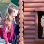 Three young girls playing at a brown wooden playhouse outdoors; two stand outside while a third peers from the window edge with a smile.