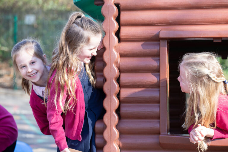 Three young girls playing at a brown wooden playhouse outdoors; two stand outside while a third peers from the window edge with a smile.