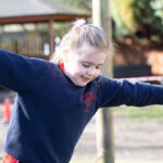 Young girl in a navy hoodie with arms outstretched at a playground, smiling.