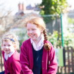 Two smiling schoolgirls in red cardigans and uniforms play outside on a sunny day in a yard with a wooden fence.