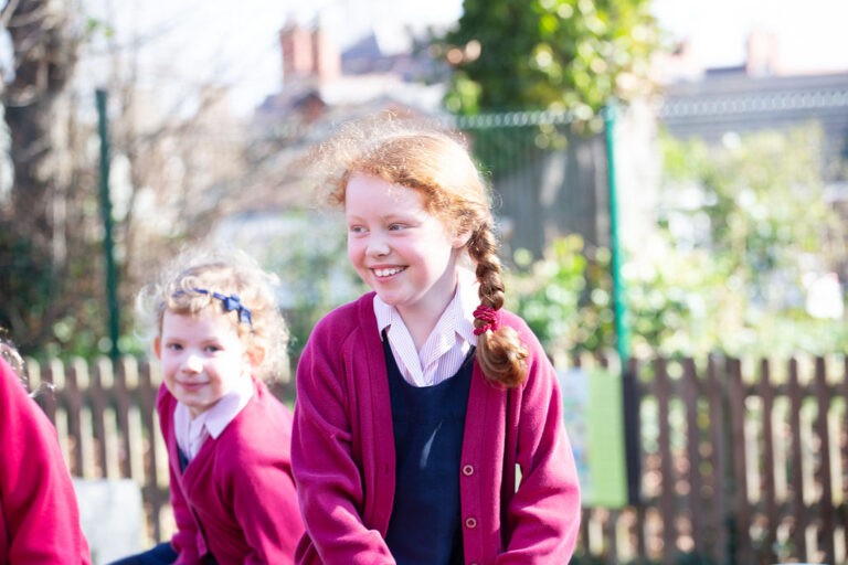 Two smiling schoolgirls in red cardigans and uniforms play outside on a sunny day in a yard with a wooden fence.