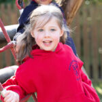 Smiling young girl in a red hoodie holding onto playground ropes outdoors, with a wooden fence in the background.