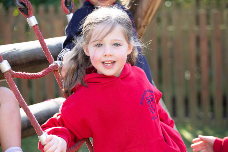 Smiling young girl in a red hoodie holding onto playground ropes outdoors, with a wooden fence in the background.