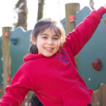 Smiling girl in a red hoodie climbs a colorful playground wall, holding onto the handgrips.