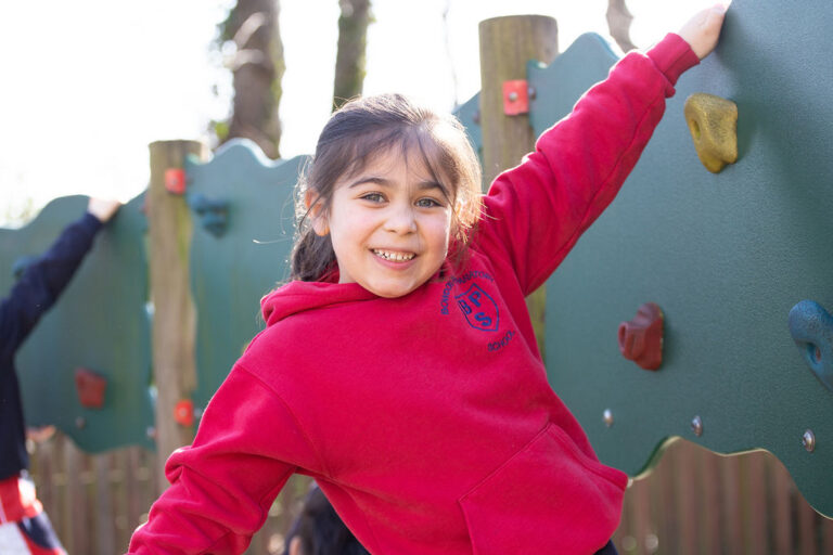Smiling girl in a red hoodie climbs a colorful playground wall, holding onto the handgrips.