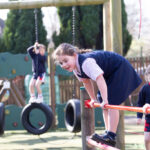 Girl in a navy uniform climbs a red horizontal bar at a playground with other kids in the background on swings and tires.