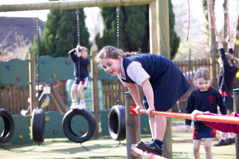 Girl in a navy uniform climbs a red horizontal bar at a playground with other kids in the background on swings and tires.