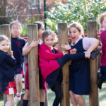 Group of smiling schoolchildren in navy and red uniforms playing on a wooden obstacle course in a sunny playground area.