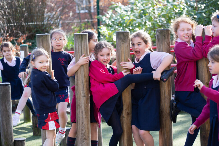 Group of smiling schoolchildren in navy and red uniforms playing on a wooden obstacle course in a sunny playground area.