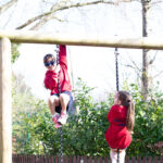 Two children on a wooden playground frame, one swinging on a rope while the other watches, in a sunny outdoor setting.