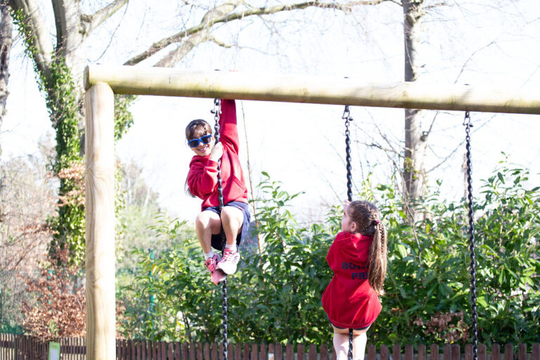 Two children on a wooden playground frame, one swinging on a rope while the other watches, in a sunny outdoor setting.