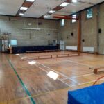Empty school gym with brick walls, wooden benches along the sides, a raised stage at the back, and blue mats in the foreground.
