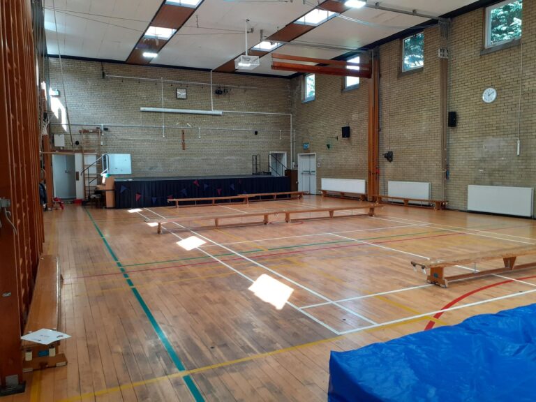 Empty school gym with brick walls, wooden benches along the sides, a raised stage at the back, and blue mats in the foreground.