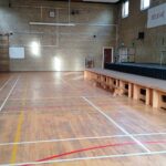 Empty school gym with a raised stage on the right, long wooden benches along the floor, and banners on the wall reading 'BELIEVE' and 'PERSEVERE'.