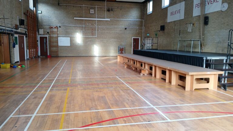 Empty school gym with a raised stage on the right, long wooden benches along the floor, and banners on the wall reading 'BELIEVE' and 'PERSEVERE'.