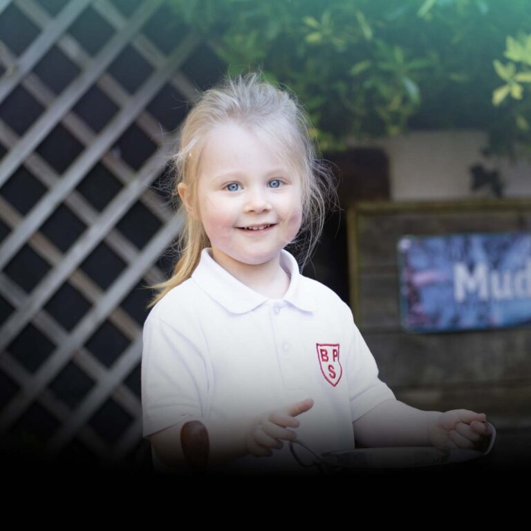 Smiling young girl with blonde hair in a white polo shirt with a crest, outdoors near a wooden trellis, holding a plate and spoon in front of her.