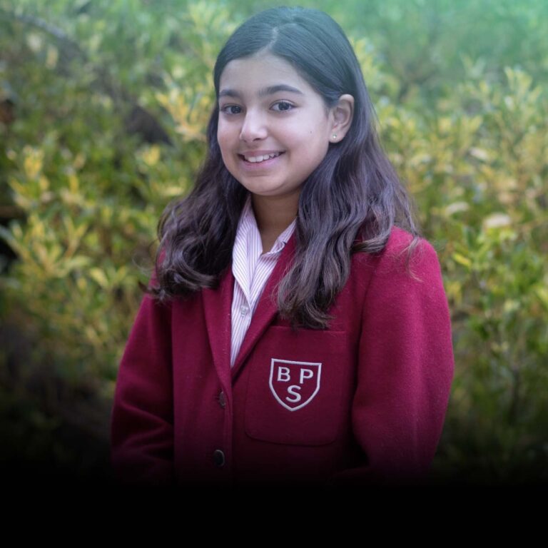 Portrait of a smiling girl wearing a maroon school blazer with a BPS crest, outdoors among green bushes.
