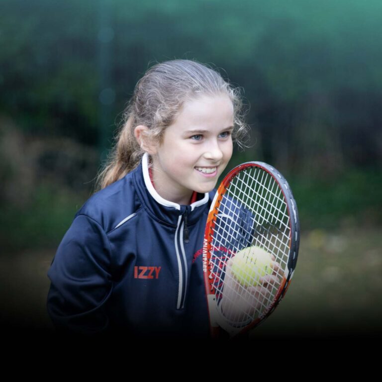 Young girl named Izzy smiling while holding a tennis racket outdoors, wearing a navy jacket with her name on it.