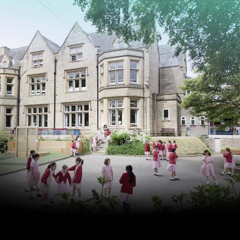 Group of schoolchildren in pink and red uniforms playing in a courtyard in front of a large stone school building with bay windows.