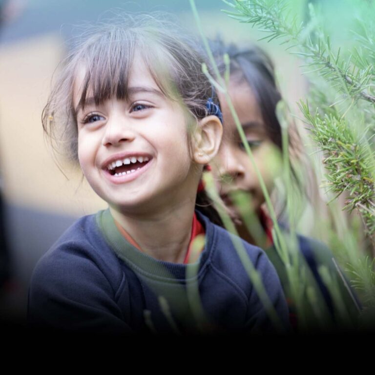 Smiling young girl with bangs outdoors among green plants, with another child partially visible in the background.