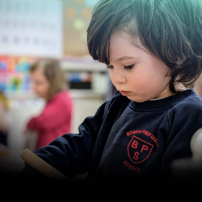 Young child in a navy sweatshirt reads a book in a classroom, with colorful posters in the blurred background.