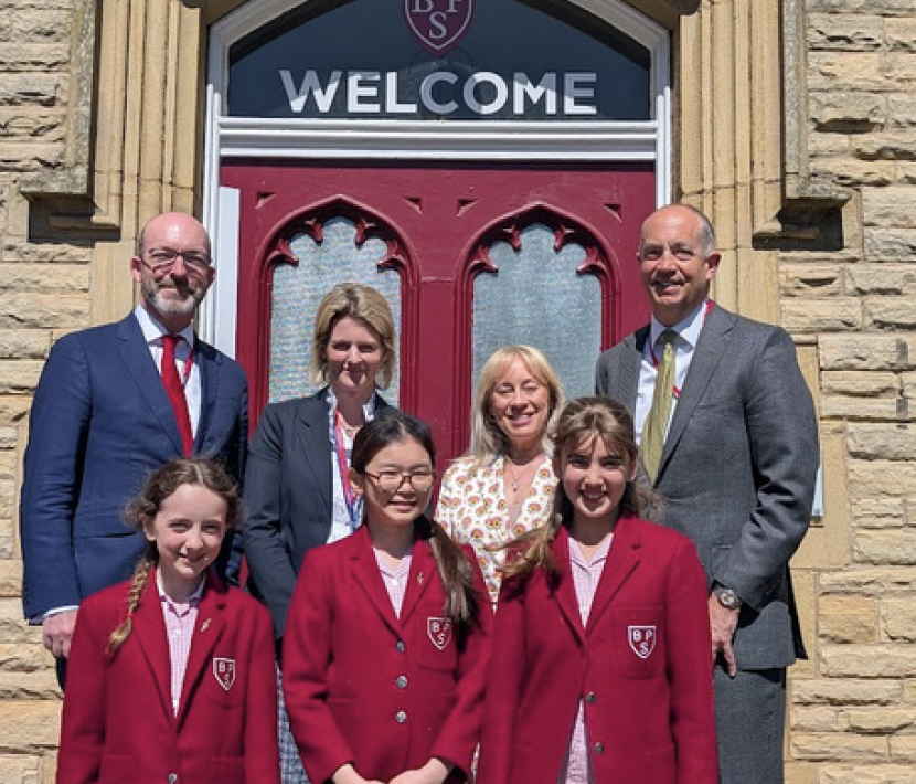 Group of students and teachers posing for a welcome photo outside a stone school with a red arched door and a 'WELCOME' sign above.
